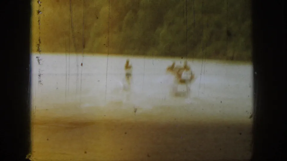 1961 Michigan lake frame with two distant figures on or near the water, seen through faded 8mm color and heavy vertical scratches.