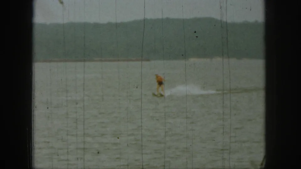 1961 Michigan 8mm frame showing a distant person water-skiing across a lake under an overcast sky.
