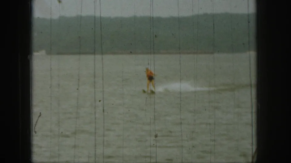 A distant skier in an orange top rides across a Michigan lake in this soft, scratched 1961 home-movie frame.