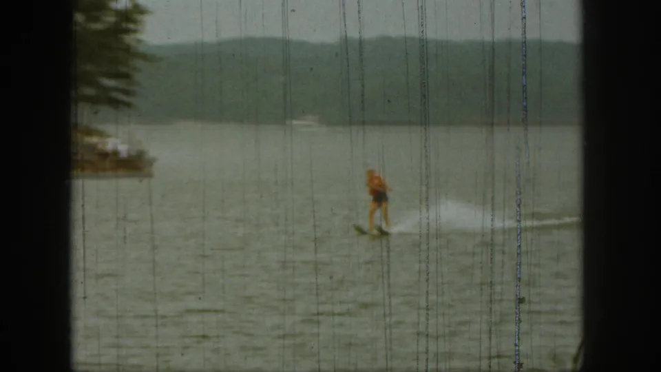 1961 Michigan home-movie still of a distant water-skier passing near a partially visible lakeshore.