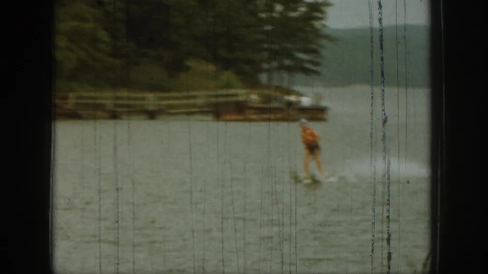 A distant skier moves across a Michigan lake near trees and a small shoreline structure in a 1961 8mm frame.
