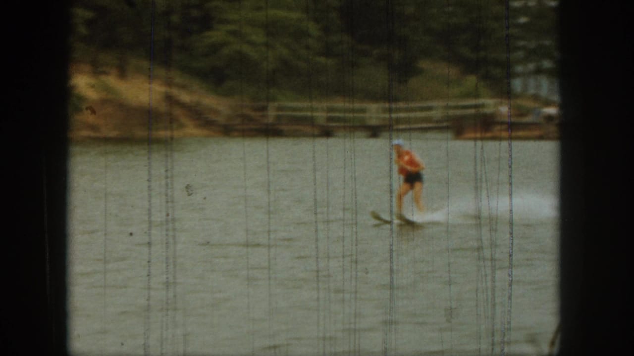 In 1961 Michigan home-movie footage, a woman water skis across a lake while trees and a low shoreline sit in the background.