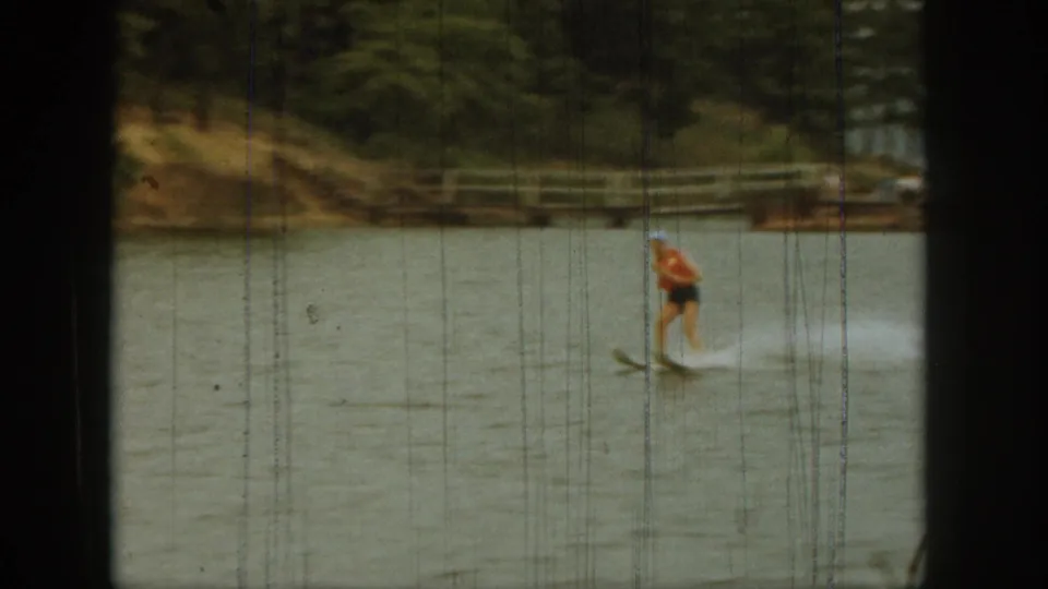 In 1961 Michigan home-movie footage, a woman water skis across a lake while trees and a low shoreline sit in the background.