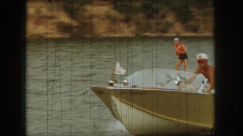A small tow boat crosses the lake as the woman water skis behind it in 1961 Michigan home-movie footage.
