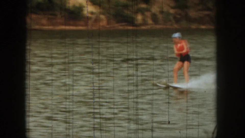 The woman, wearing a red life vest and light swim cap, skis closer across the Michigan lake in this 1961 8mm frame.