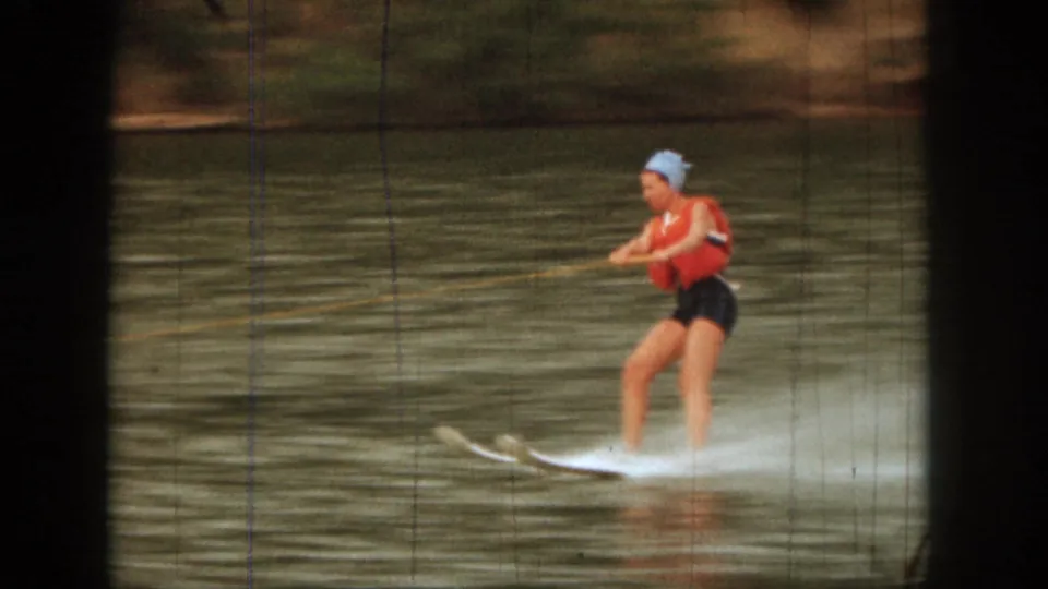 A closer 1961 Michigan home-movie frame shows the woman skiing across the lake in a red life vest and light blue swim cap.