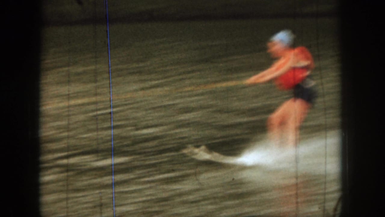 1961 Michigan home-movie frame showing a water skier in a red life vest holding a tow rope while spray rises from the lake.