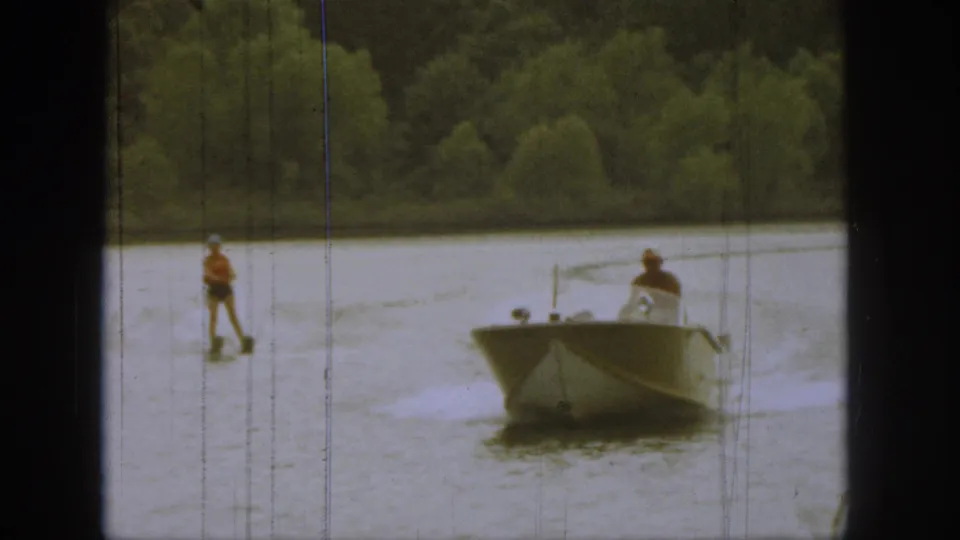 1961 Michigan home-movie view of a skier trailing a small motorboat on a lake with trees in the background.