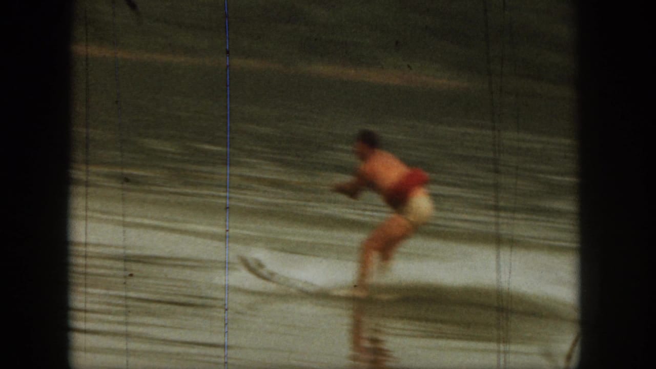 1961 Michigan 8mm frame showing a man in red swimwear water skiing on a lake, with blurred motion and tree-lined shore behind him.