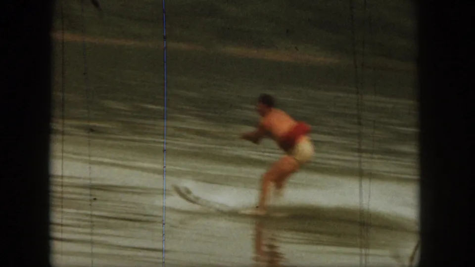 1961 Michigan 8mm frame showing a man in red swimwear water skiing on a lake, with blurred motion and tree-lined shore behind him.