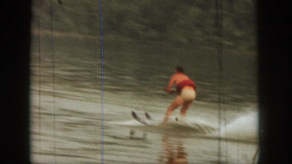 1961 Michigan home-movie frame of a man water skiing away from the camera, leaving a white wake on a wooded lake.