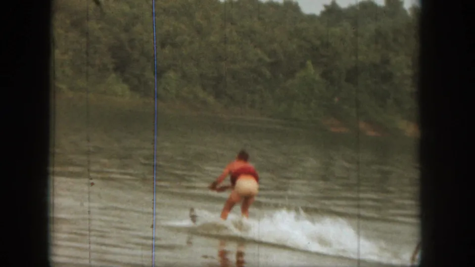 1961 Michigan 8mm frame showing the skier moving farther from the camera across a lake bordered by dense trees.