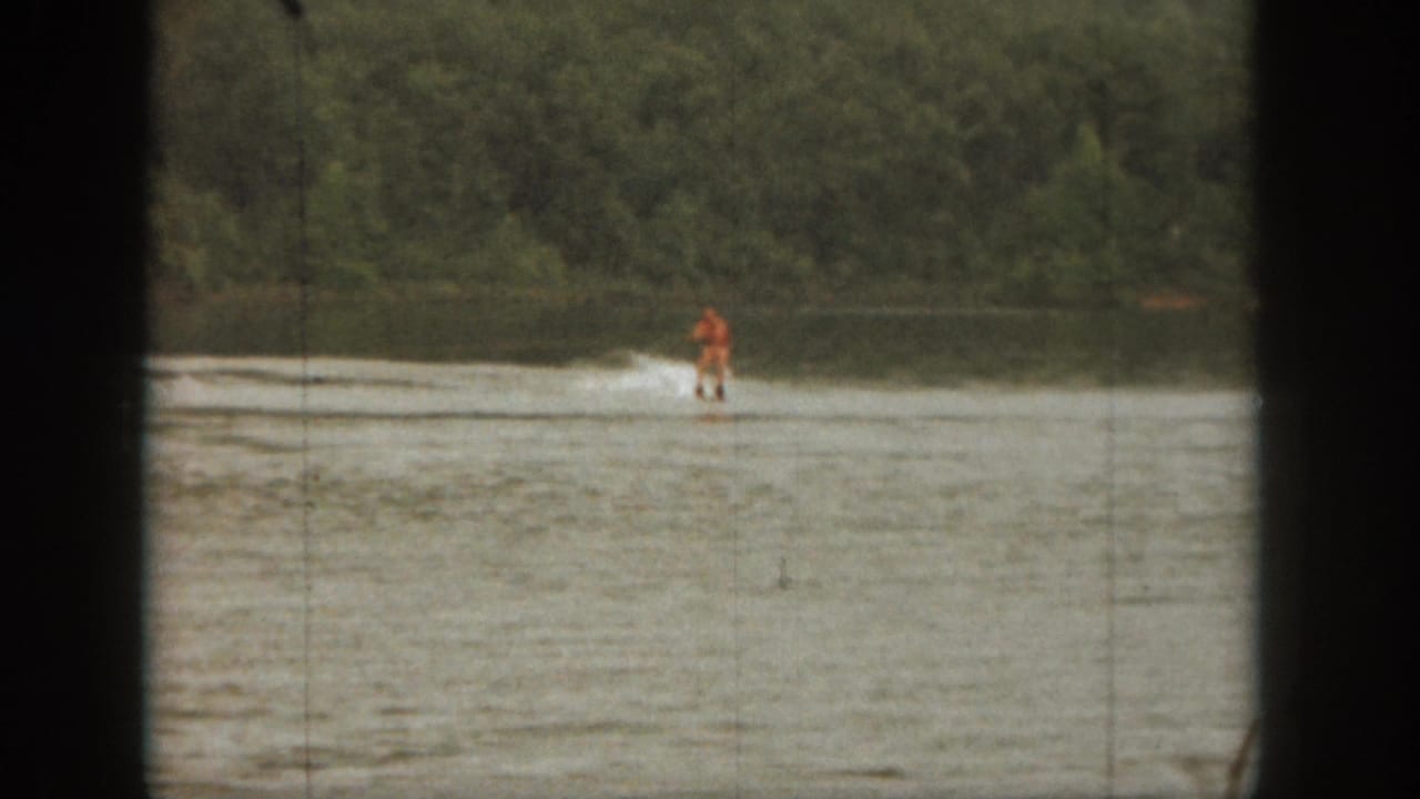 Distant water skier in orange crossing a lake near a wooded shoreline.