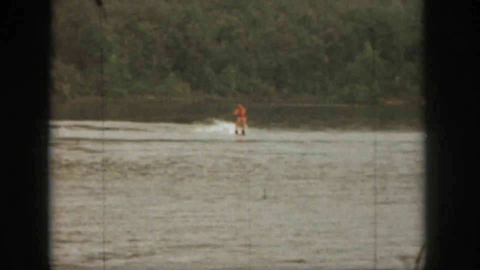 Distant water skier in orange gliding across a Michigan lake in 1961 home-movie footage, with trees lining the far shore.