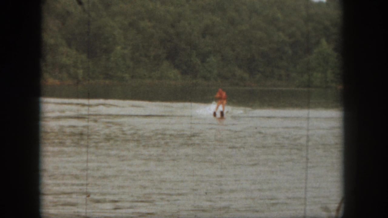Water skier in orange near the far side of a calm lake.