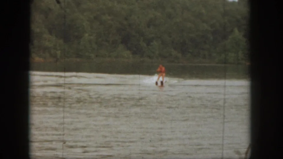 A small figure in orange skis across the water near a tree-lined Michigan shoreline in vintage 8mm footage from 1961.