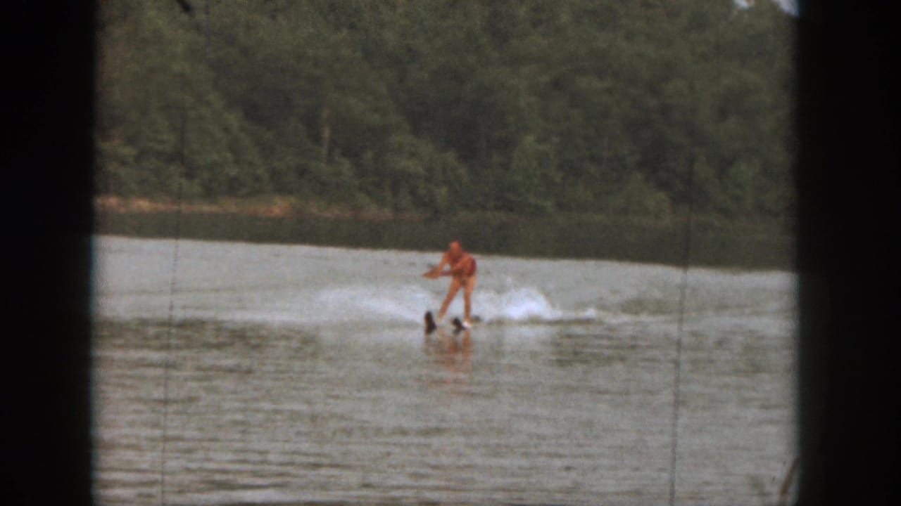 1961 Michigan 8mm frame showing a distant water skier in orange crossing a calm lake with trees along the far shore.