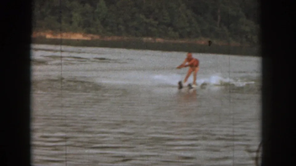 1961 Michigan home-movie frame with a distant skier in orange moving across open lake water near a tree-lined shore.