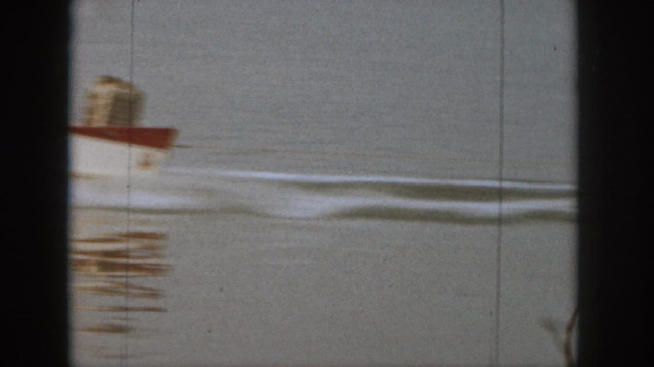 1961 Michigan home-movie frame showing a small red-and-white motorboat moving across calm lake water with a trailing wake.