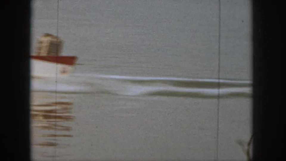 1961 Michigan home-movie frame showing a small red-and-white motorboat moving across calm lake water with a trailing wake.