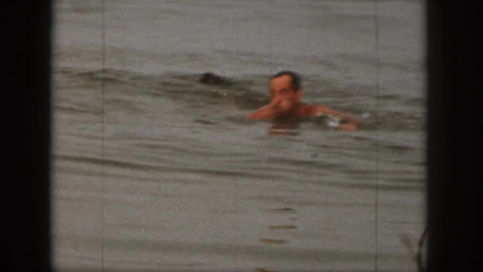 Adult man swims through the lake after the waterski fall, filmed in soft-focus 8mm home-movie style in Michigan in 1961.