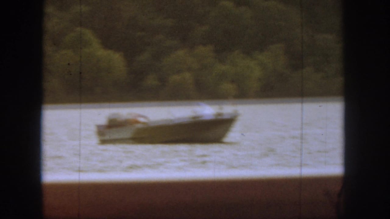 A small motorboat sits on the Michigan lake as the swimmer returns, with wooded shoreline and 8mm film softness visible.