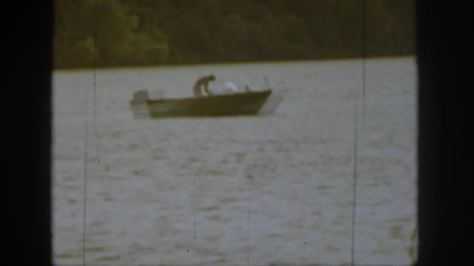 1961 Michigan 8mm frame showing a man bending inside a small boat on rippling lake water with trees in the distance.
