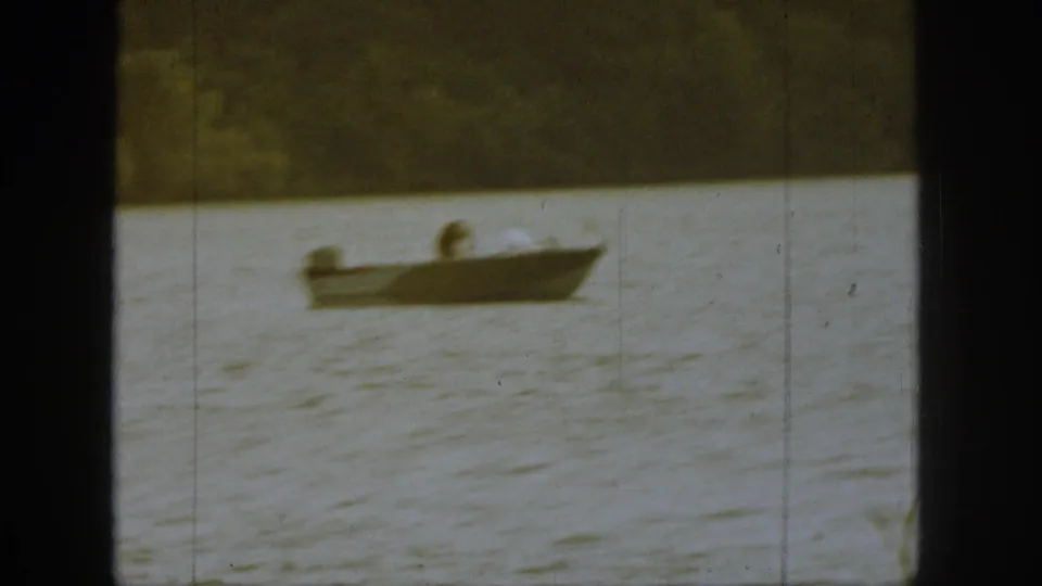 1961 Michigan home-movie frame of a small boat on lake water, with an adult figure low in the boat and wooded shoreline behind.