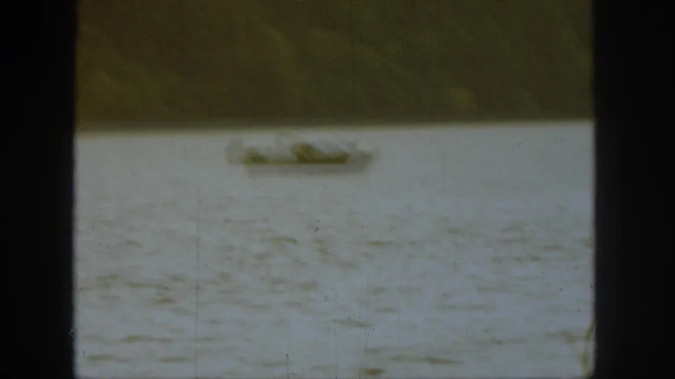 Distant 1961 Michigan lake frame with a small boat and adult figure softened by grain, water texture, and dark film borders.