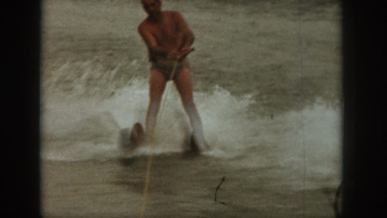 1961 Michigan home-movie frame of an adult man water skiing on a lake, with white spray and soft 8mm grain.