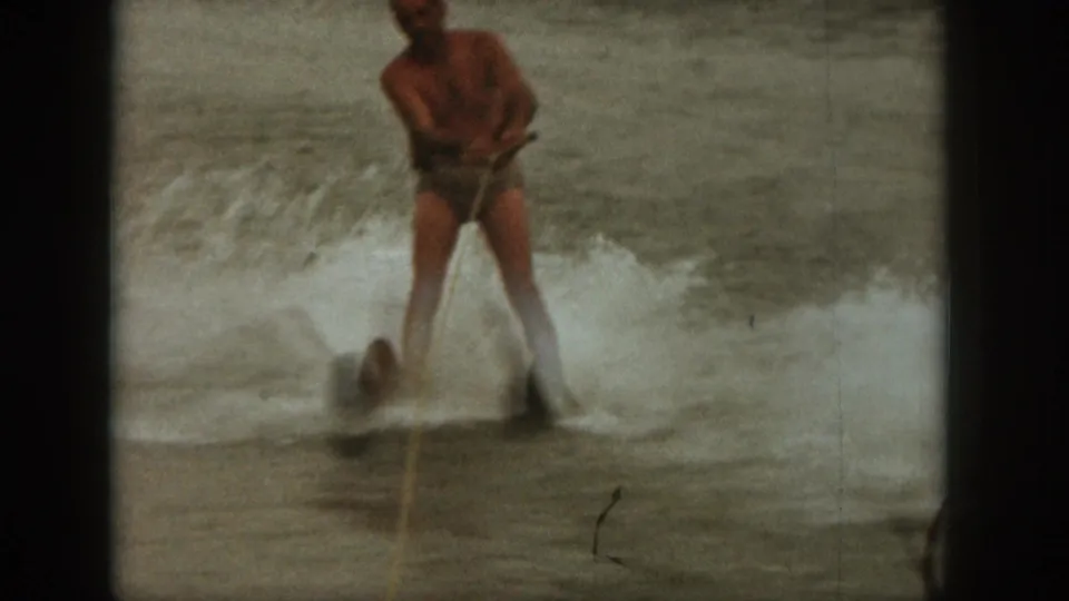 1961 Michigan home-movie frame of an adult man water skiing on a lake, with white spray and soft 8mm grain.