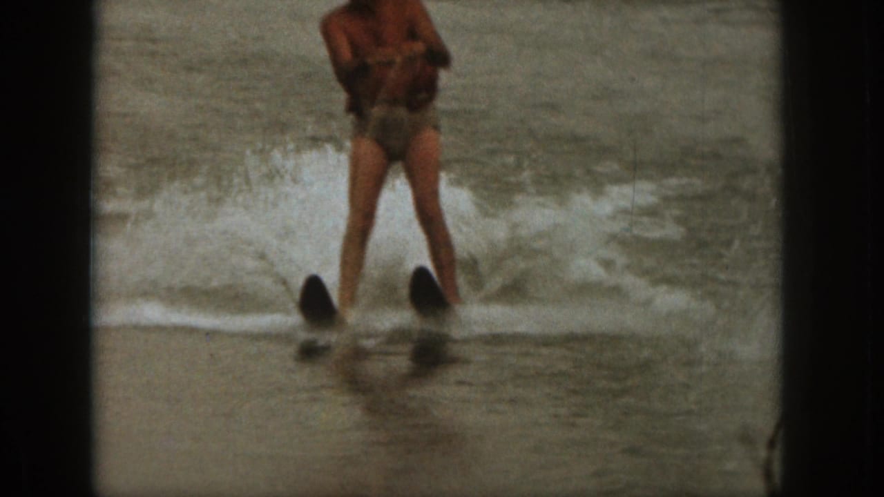 Adult man on water skis faces the camera as waves break behind him.