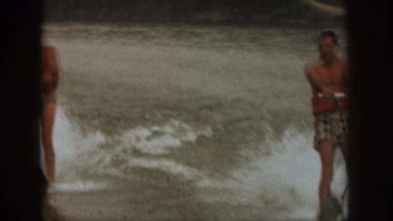 1961 Michigan home-movie frame with two adult men near the water, one partly cropped and one in a red life jacket.