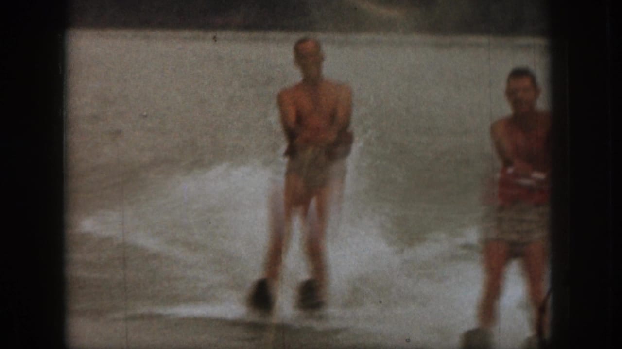 1961 Michigan 8mm frame showing two adult men in the lake, one water skiing and another wearing a red life jacket.