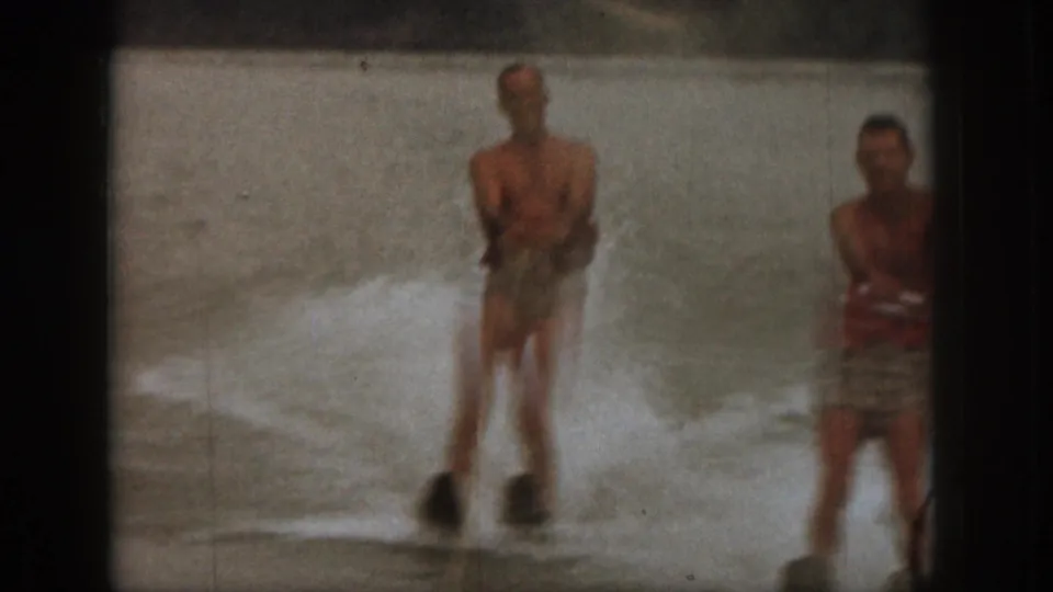 1961 Michigan 8mm frame showing two adult men in the lake, one water skiing and another wearing a red life jacket.