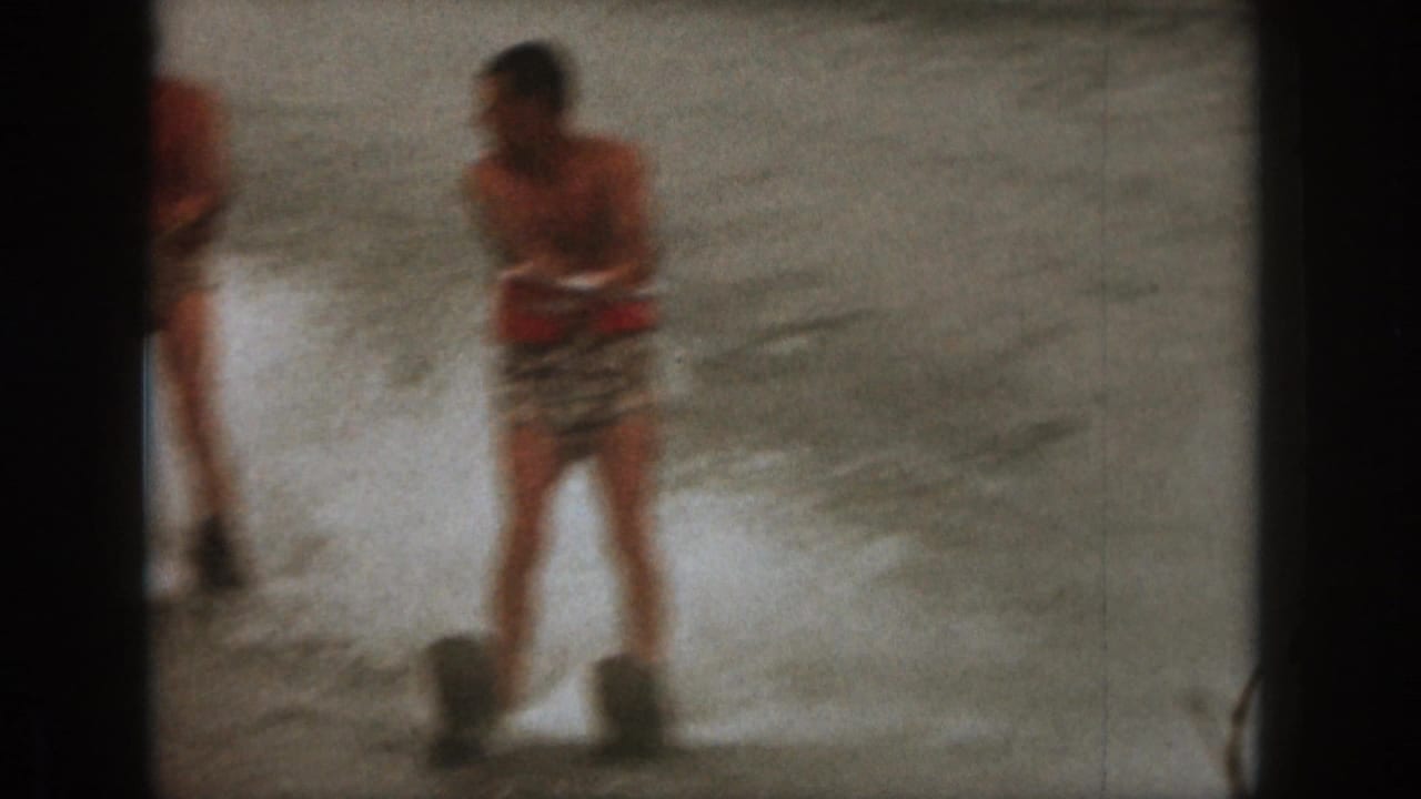 1961 Michigan home-movie frame showing an adult man water skiing through spray in patterned trunks and a red life vest.