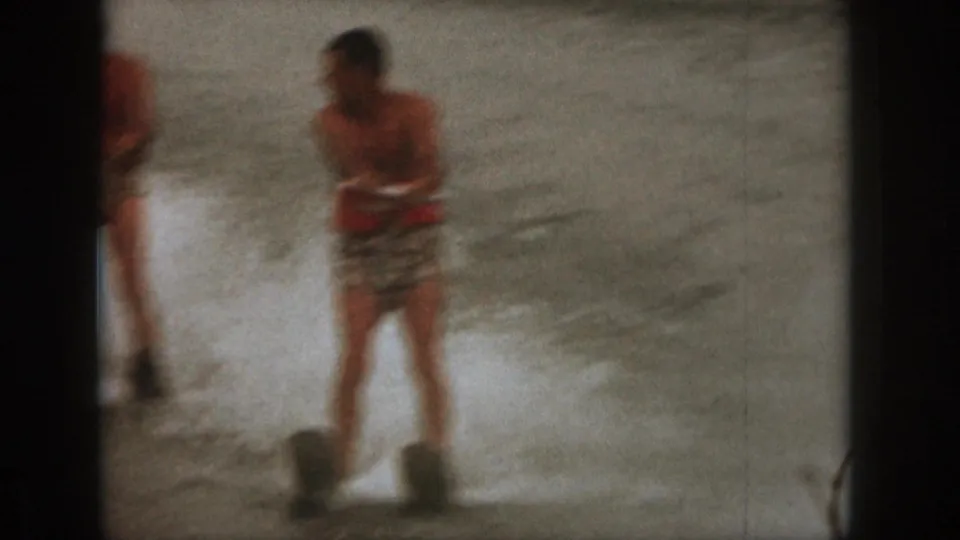 1961 Michigan home-movie frame showing an adult man water skiing through spray in patterned trunks and a red life vest.