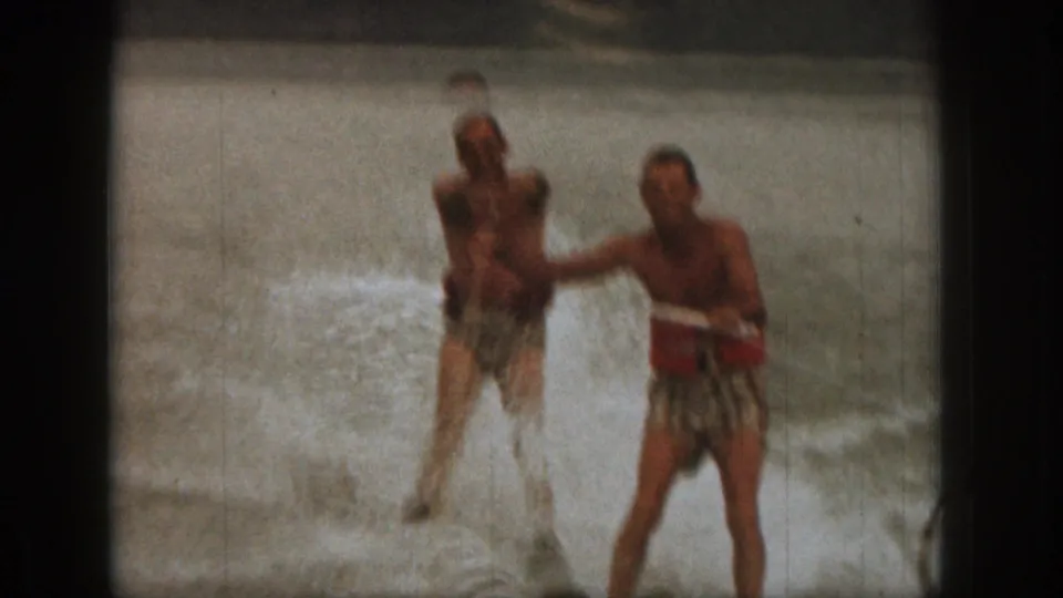 1961 Michigan home-movie frame with two adult men water skiing together, one in a red life vest, amid white spray.