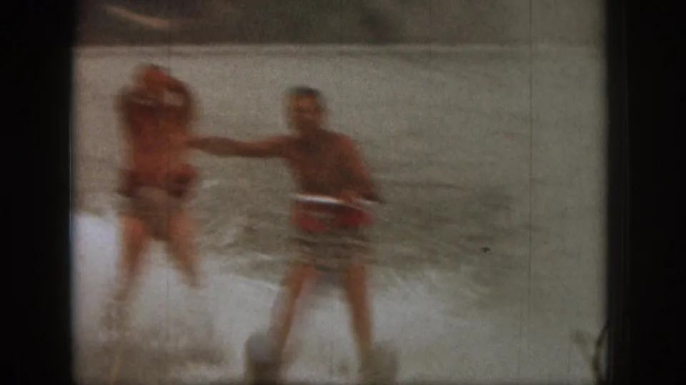 Soft 1961 Michigan home-movie frame showing two adult men water skiing, with one arm extended as spray rises around them.