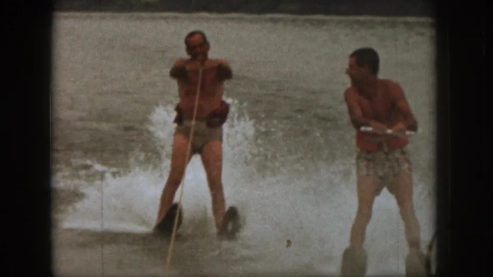 1961 Michigan home-movie frame of two adult men water skiing, with a visible tow rope and lake spray behind them.