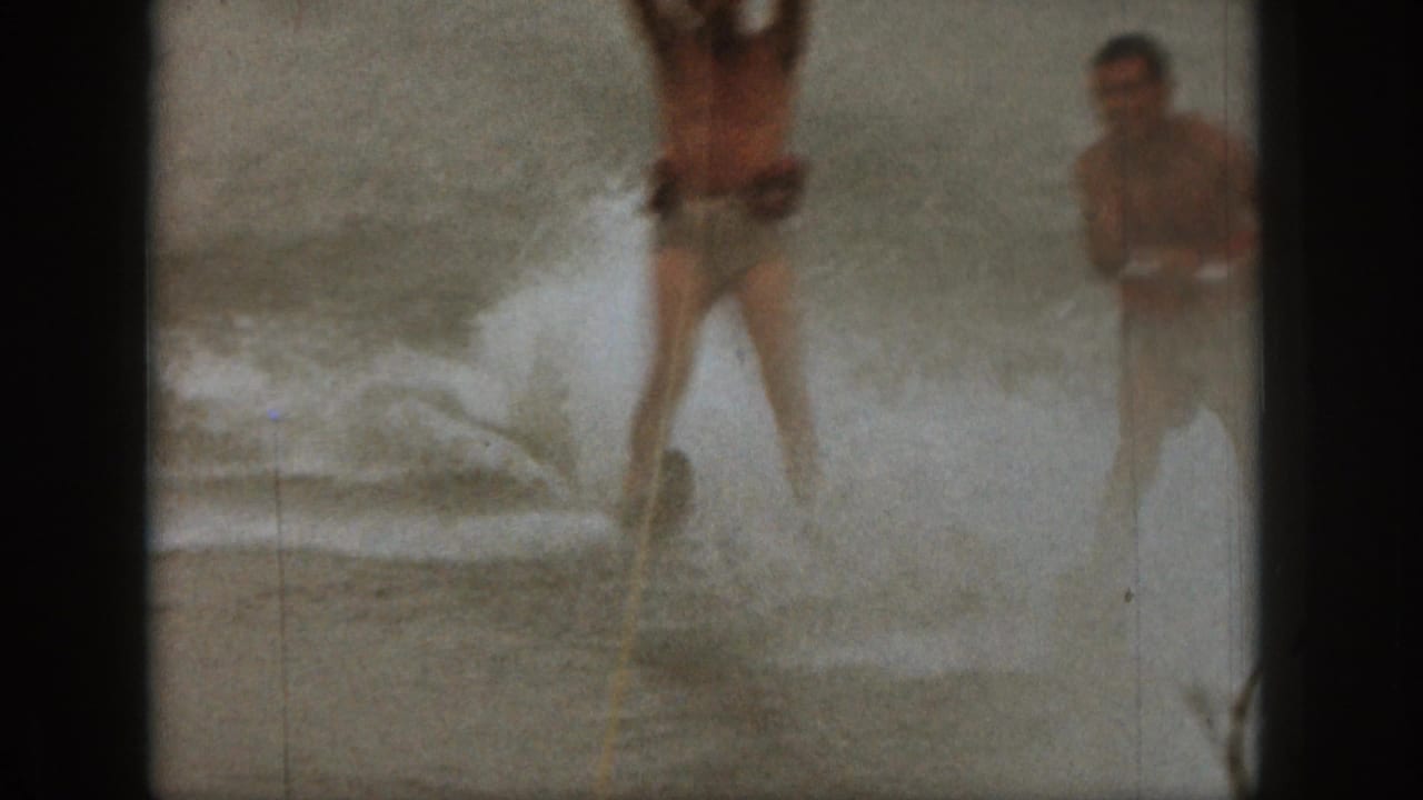 1961 Michigan home-movie frame showing two men water skiing across choppy lake water, partly obscured by spray and film softness.