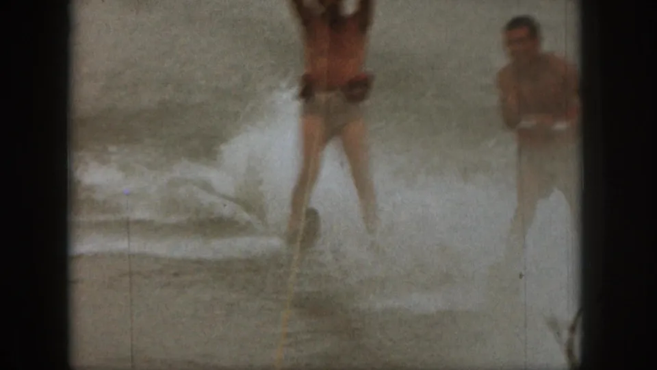 1961 Michigan home-movie frame showing two men water skiing across choppy lake water, partly obscured by spray and film softness.