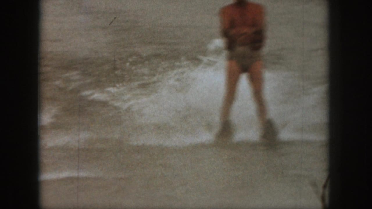 1961 Michigan 8mm frame showing an adult water skier in swim trunks holding a tow handle amid splashing lake water.