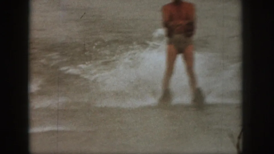 1961 Michigan 8mm frame showing an adult water skier in swim trunks holding a tow handle amid splashing lake water.