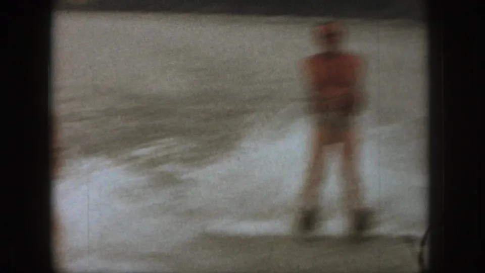 Soft 1961 Michigan home-movie frame of an adult water skier being pulled through shallow lake water.