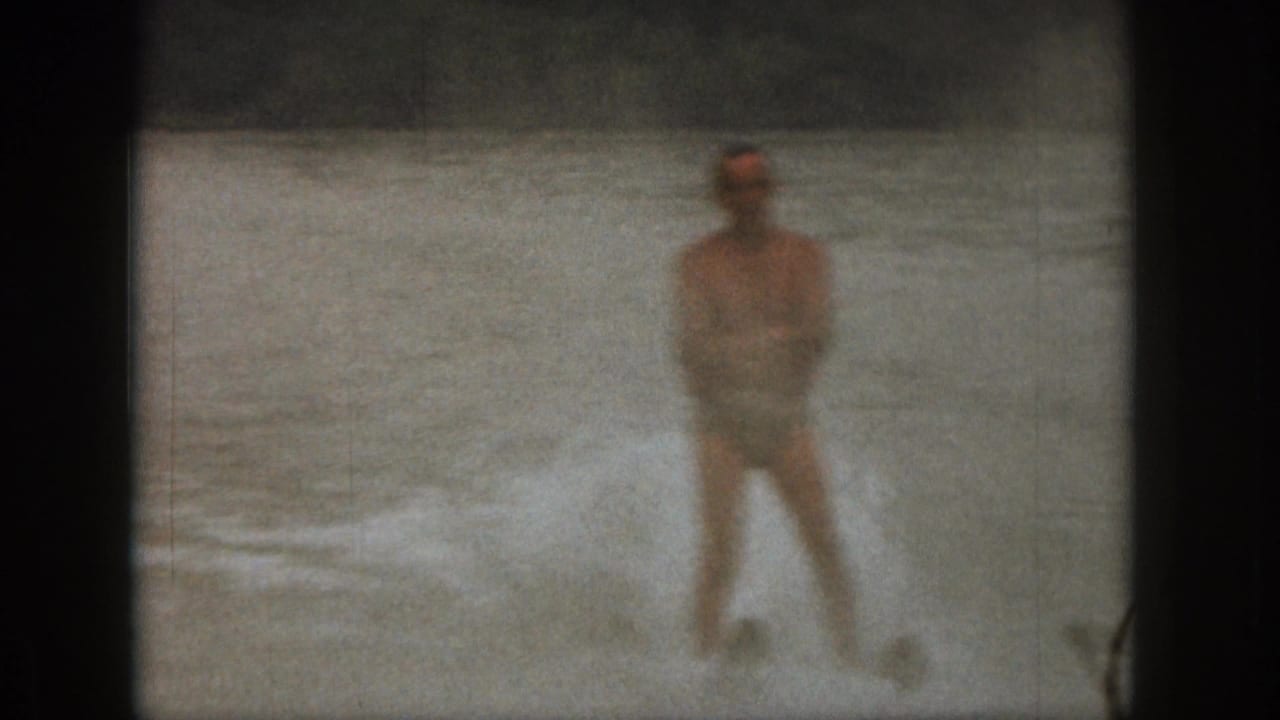 Adult man water skiing on a Michigan lake in 1961 home-movie footage, with spray around his legs and a distant shoreline.