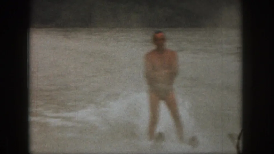 Adult man water skiing on a Michigan lake in 1961 home-movie footage, with spray around his legs and a distant shoreline.