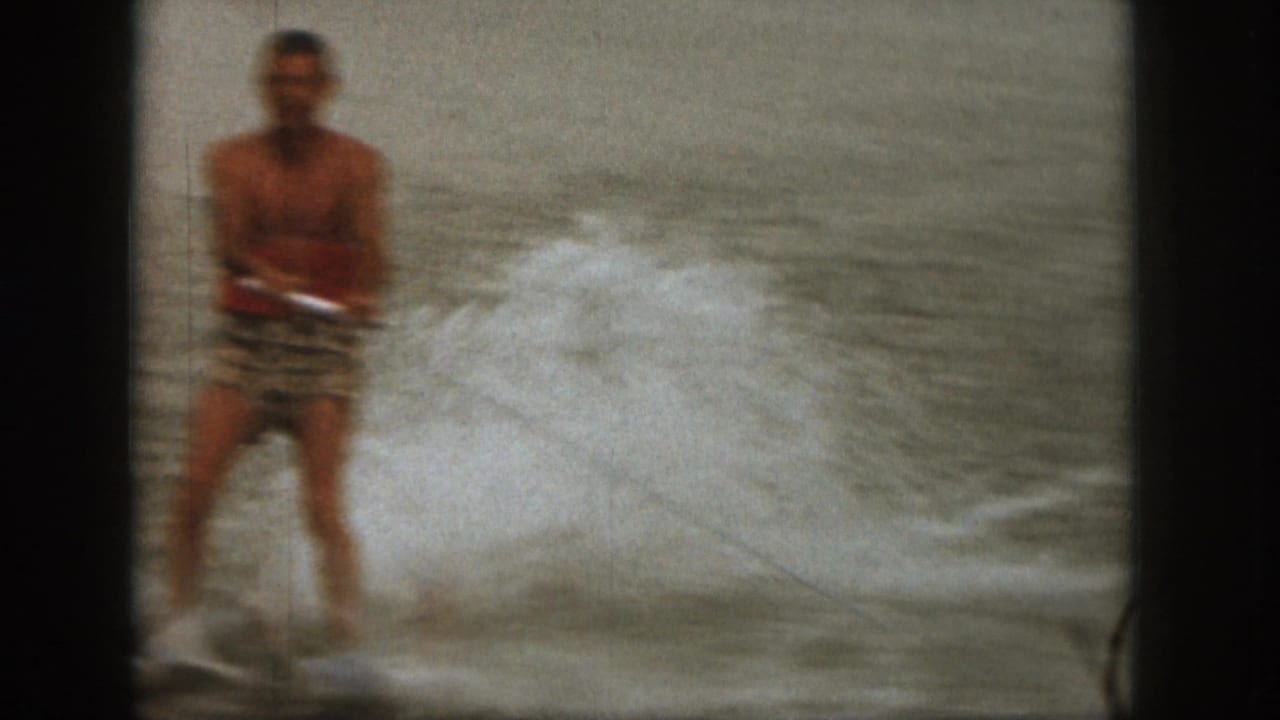 The skier appears closer to the camera, facing forward in swim trunks as lake spray breaks beside him in vintage 8mm footage.