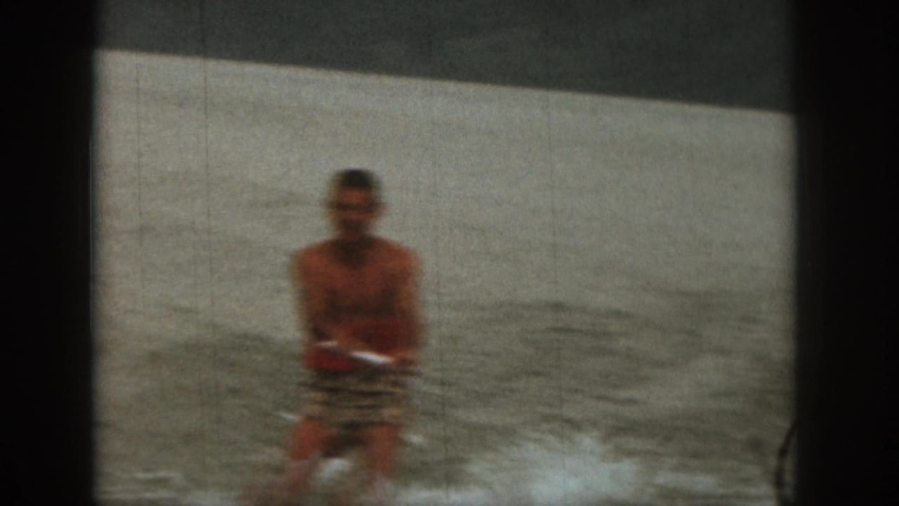An adult water skier continues across the Michigan lake, framed by muted 8mm color and a pale shoreline in the background.