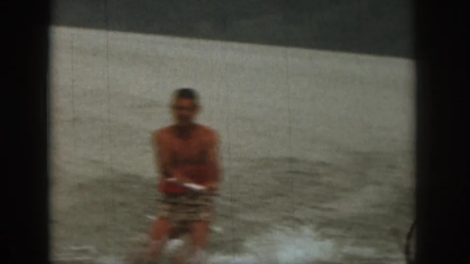 An adult water skier continues across the Michigan lake, framed by muted 8mm color and a pale shoreline in the background.